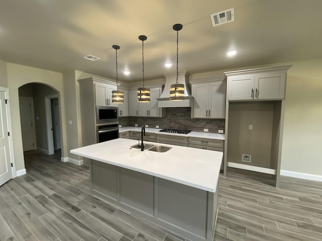 kitchen with custom exhaust hood, a center island with sink, appliances with stainless steel finishes, tasteful backsplash, and decorative light fixtures