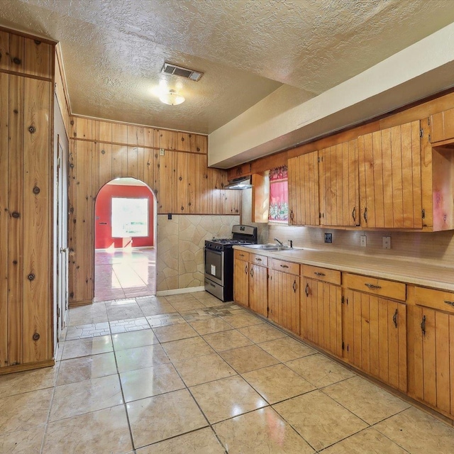 1710 17th St, Port Arthur, TX 77640 - kitchen featuring brown cabinets, light countertops, arched walkways, wood walls, and stainless steel range with gas cooktop
