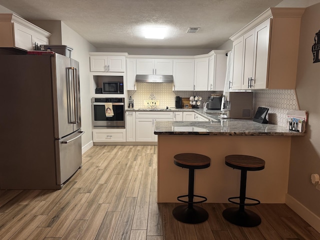 kitchen featuring dark stone countertops, light wood-style flooring, stainless steel appliances, under cabinet range hood, and a peninsula