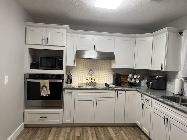 kitchen featuring white cabinets, under cabinet range hood, oven, wood finished floors, and backsplash