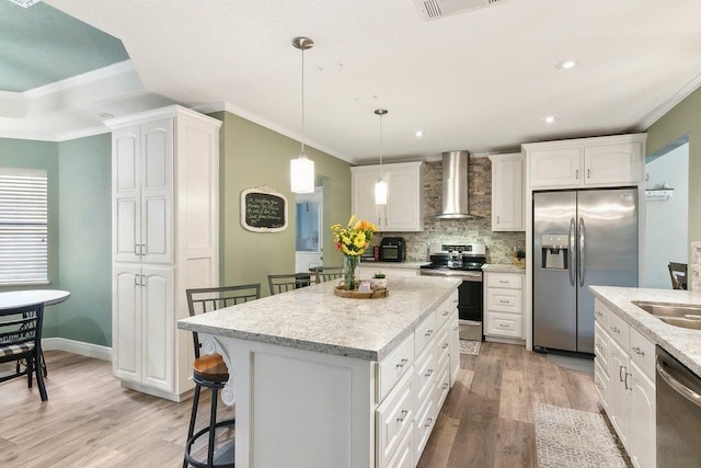 kitchen featuring wall chimney exhaust hood, stainless steel appliances, white cabinets, a kitchen island, and hanging light fixtures