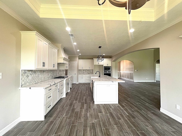 kitchen with arched walkways, premium range hood, a center island with sink, and white cabinetry