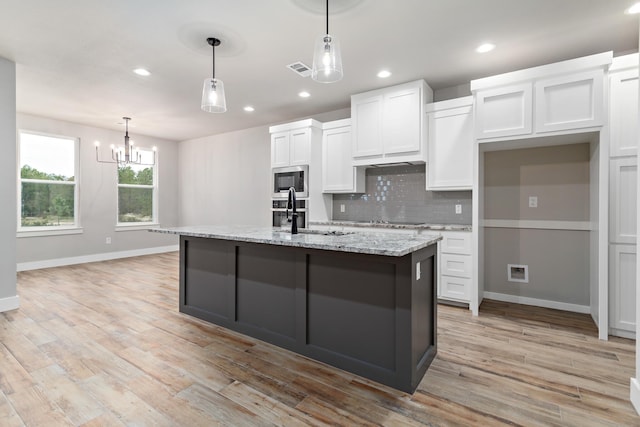 kitchen with stainless steel microwave, a kitchen island with sink, sink, light stone countertops, and white cabinetry