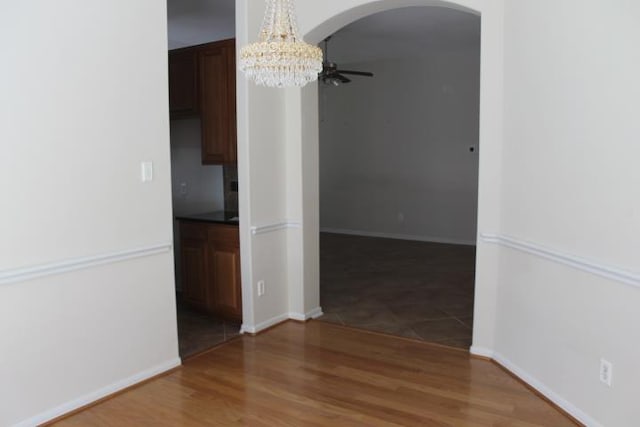 unfurnished dining area with ceiling fan with notable chandelier, wood finished floors, arched walkways, and baseboards