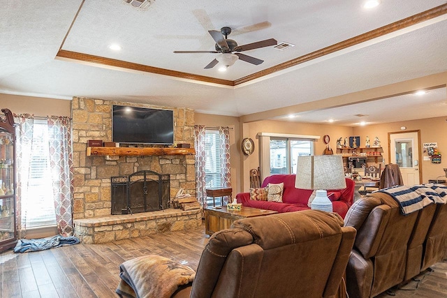 living room featuring a raised ceiling, a stone fireplace, crown molding, and wood-type flooring