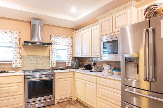 kitchen with wall chimney exhaust hood, premium appliances, backsplash, and light hardwood / wood-style flooring