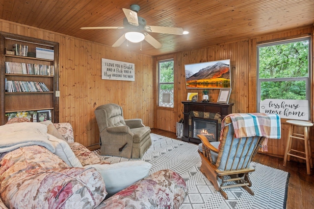 sitting room with wood walls, a stone fireplace, wood finished floors, a ceiling fan, and wooden ceiling