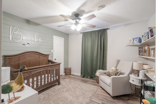bedroom with a textured ceiling, a ceiling fan, a nursery area, and wood finished floors