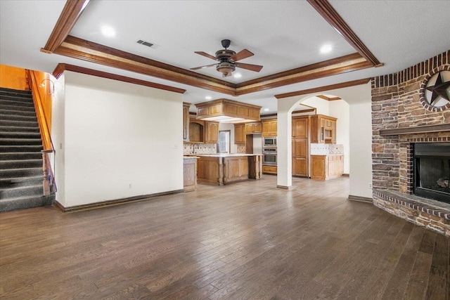 unfurnished living room featuring a stone fireplace, ceiling fan, ornamental molding, a tray ceiling, and dark hardwood / wood-style flooring