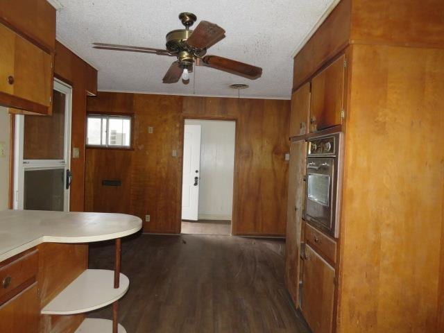 kitchen with a textured ceiling, ceiling fan, dark wood-type flooring, oven, and wood walls