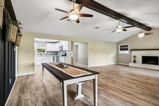 kitchen with decorative backsplash, appliances with stainless steel finishes, a brick fireplace, lofted ceiling with beams, and white cabinetry