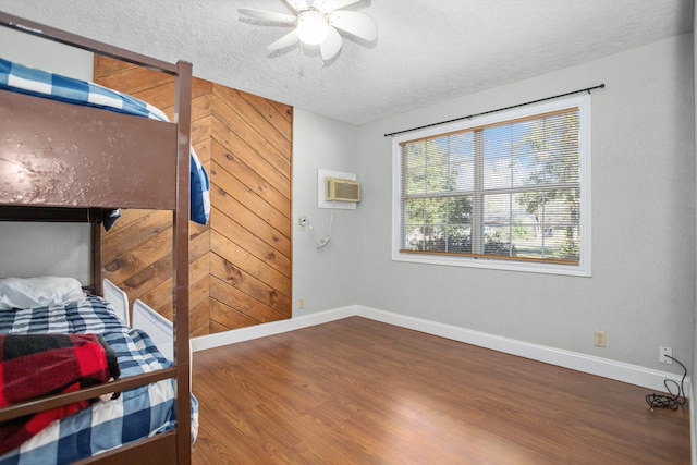 bedroom featuring an AC wall unit, wooden walls, hardwood / wood-style flooring, ceiling fan, and a textured ceiling