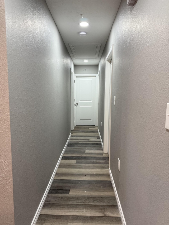 hall featuring a textured wall, dark wood finished floors, attic access, and baseboards