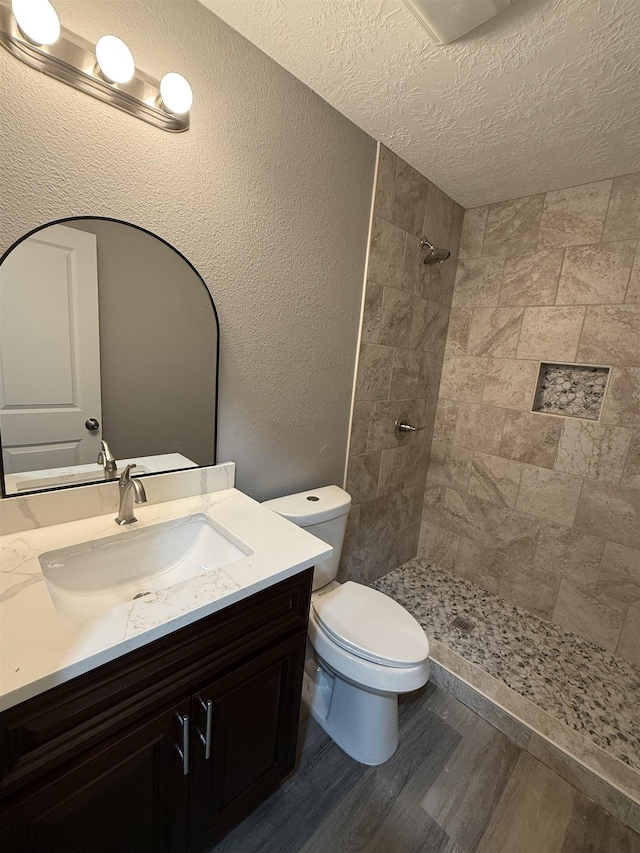 bathroom featuring a textured wall, toilet, wood finished floors, vanity, and tiled shower