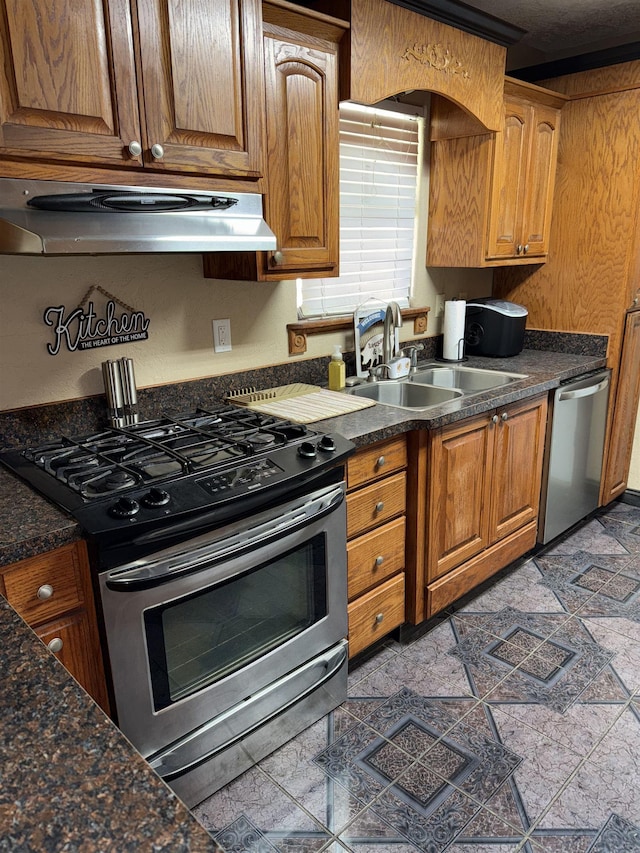 kitchen featuring dark countertops, under cabinet range hood, appliances with stainless steel finishes, brown cabinetry, and a sink