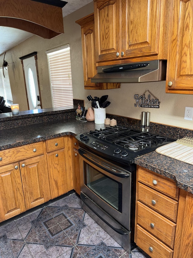 kitchen featuring under cabinet range hood, arched walkways, a peninsula, brown cabinetry, and stainless steel range with gas stovetop