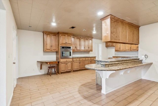 kitchen with kitchen peninsula, tasteful backsplash, light stone counters, oven, and a breakfast bar area