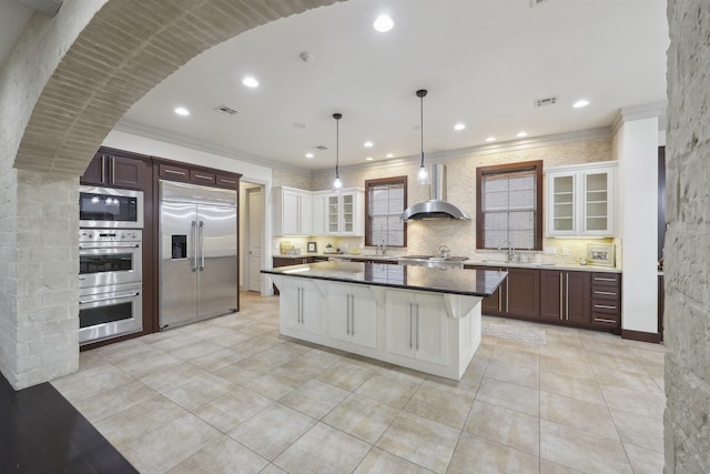 kitchen with built in appliances, glass insert cabinets, visible vents, backsplash, and wall chimney range hood