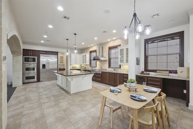 kitchen featuring a sink, built in appliances, glass insert cabinets, visible vents, and wall chimney range hood