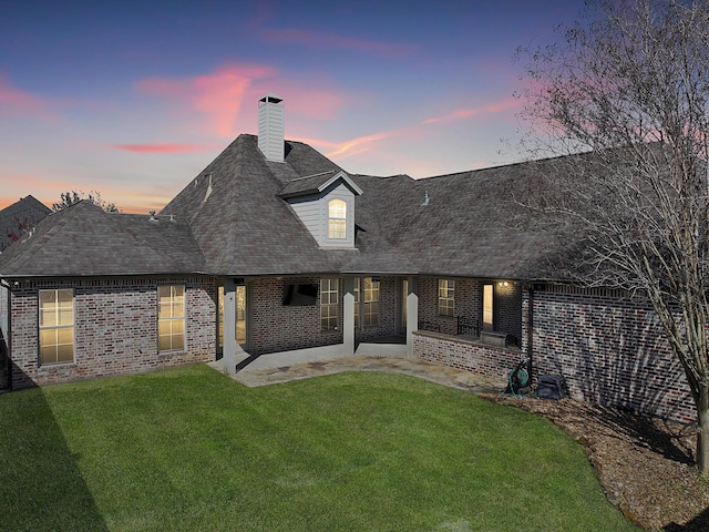 back of property at dusk with roof with shingles, a chimney, a yard, and brick siding