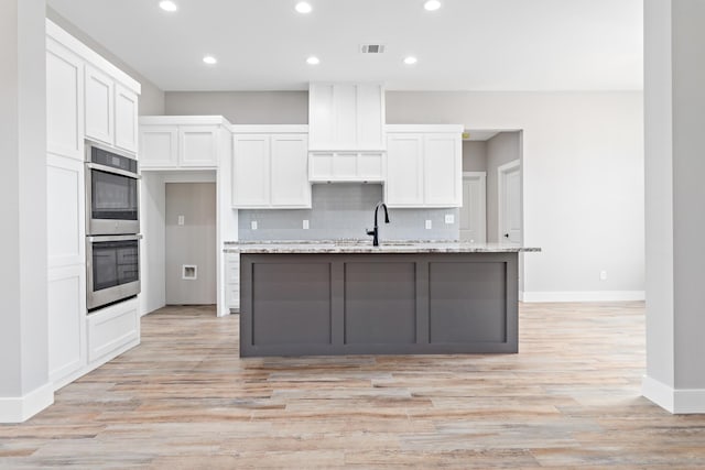 kitchen with light stone countertops, double oven, a kitchen island with sink, light hardwood / wood-style floors, and white cabinetry