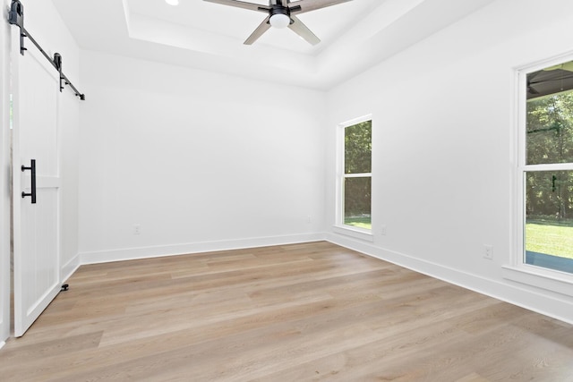 unfurnished room with ceiling fan, a barn door, light hardwood / wood-style flooring, and a tray ceiling