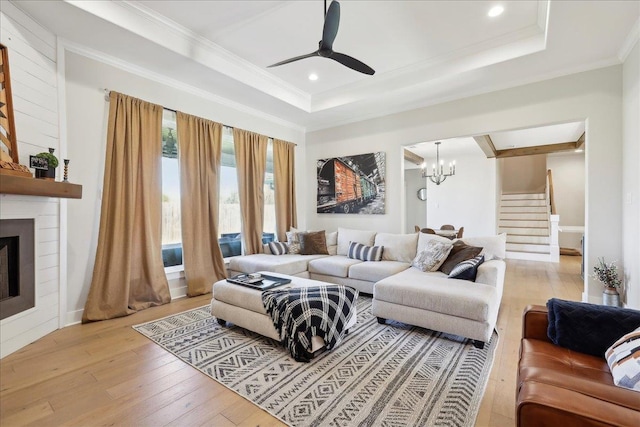 living room featuring a tray ceiling, a large fireplace, light wood-style floors, crown molding, and stairs