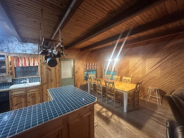 kitchen featuring beam ceiling, black dishwasher, wood finished floors, wood walls, and tile countertops