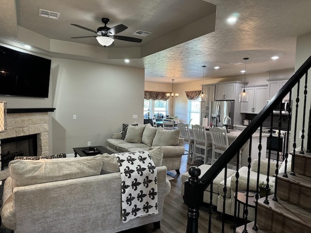 living room with a textured ceiling, a stone fireplace, dark hardwood / wood-style flooring, and ceiling fan with notable chandelier