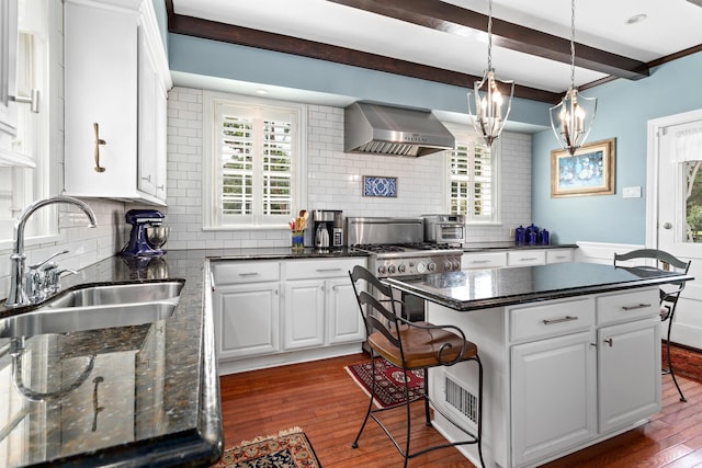 kitchen with sink, wall chimney range hood, beamed ceiling, white cabinets, and a kitchen island