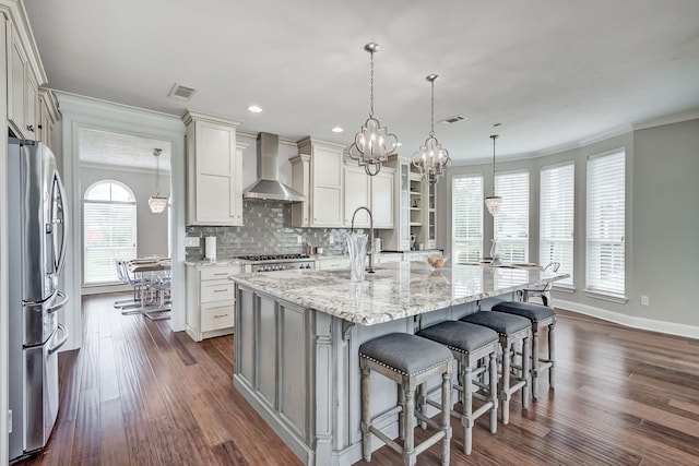 kitchen featuring stainless steel fridge, wall chimney exhaust hood, crown molding, hanging light fixtures, and an island with sink