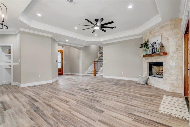 unfurnished living room featuring ceiling fan with notable chandelier, a stone fireplace, crown molding, a tray ceiling, and light hardwood / wood-style floors