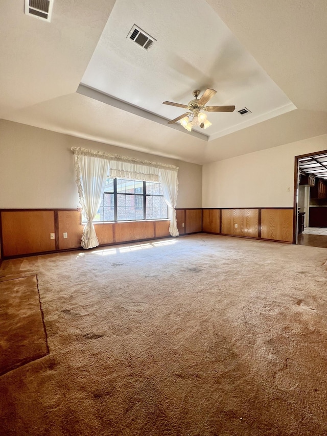 carpeted empty room featuring a raised ceiling, ceiling fan, and wooden walls