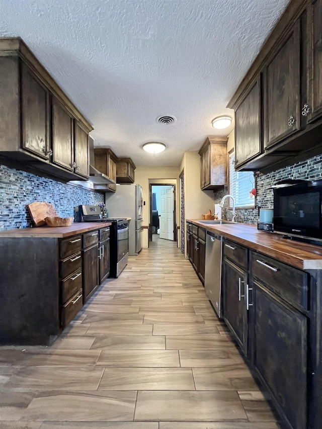 kitchen with dark brown cabinetry, butcher block counters, sink, stainless steel appliances, and tasteful backsplash