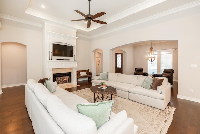 living room with dark wood-type flooring, ornamental molding, ceiling fan with notable chandelier, and a raised ceiling