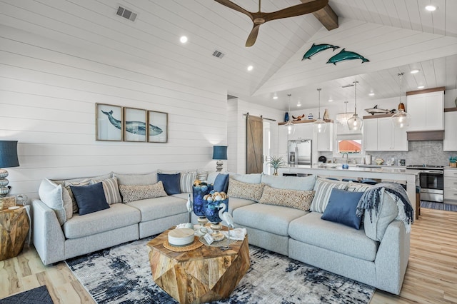 living room with beam ceiling, ceiling fan, a barn door, high vaulted ceiling, and light hardwood / wood-style floors