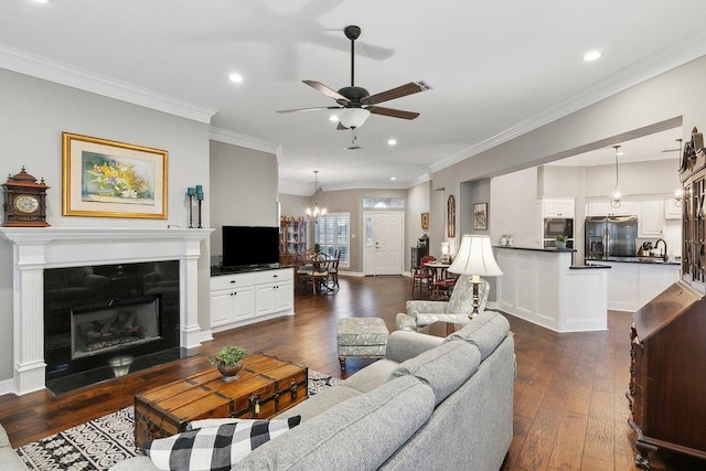 living room featuring ceiling fan with notable chandelier, dark wood-type flooring, a fireplace, sink, and crown molding