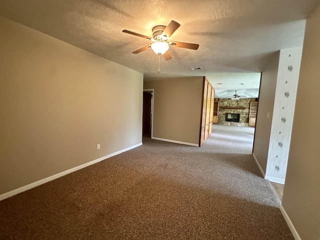 carpeted empty room with visible vents, a textured ceiling, a stone fireplace, baseboards, and ceiling fan