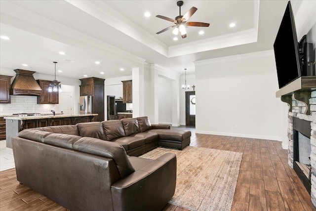 living room with wood finished floors, a fireplace, crown molding, a raised ceiling, and ceiling fan with notable chandelier