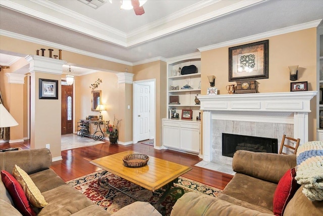 living room with ornate columns, wood finished floors, a ceiling fan, a tiled fireplace, and built in shelves