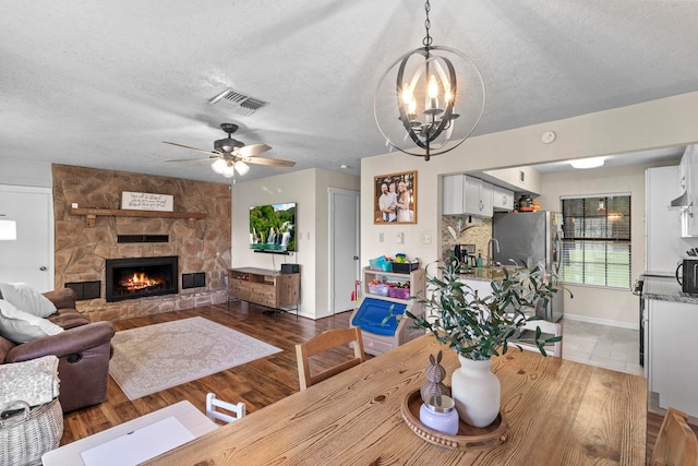 dining space featuring visible vents, a textured ceiling, a stone fireplace, and ceiling fan with notable chandelier