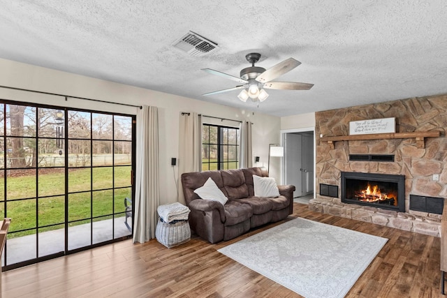 living room featuring visible vents, a textured ceiling, wood finished floors, a stone fireplace, and ceiling fan