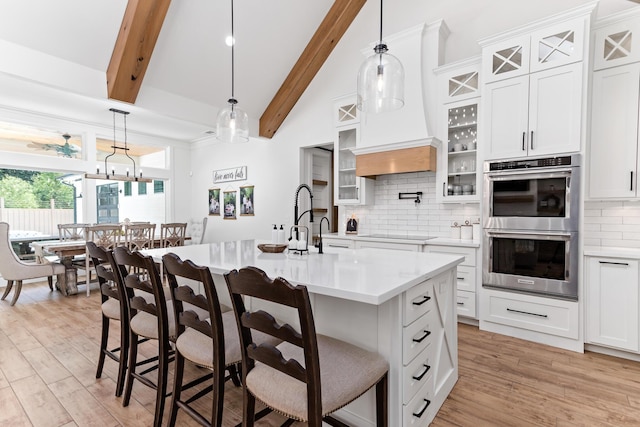 kitchen with a breakfast bar, stainless steel double oven, beam ceiling, white cabinetry, and an island with sink