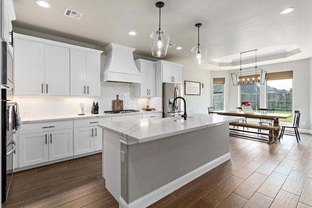 kitchen featuring custom exhaust hood, decorative light fixtures, a center island with sink, a raised ceiling, and white cabinets