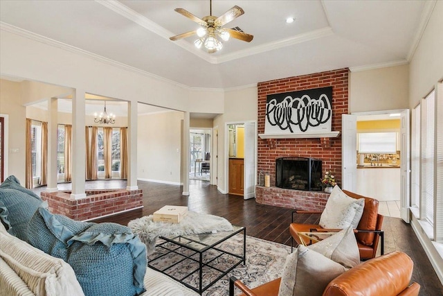 living room with a raised ceiling, crown molding, dark wood-type flooring, a fireplace, and ceiling fan with notable chandelier