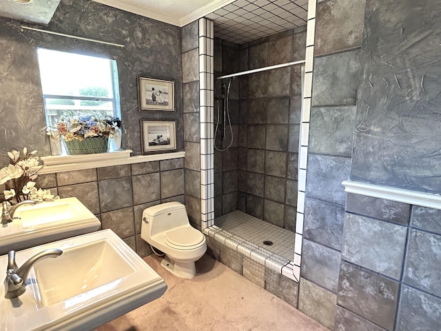 bathroom featuring a tile shower, crown molding, toilet, dual sinks, and tile walls