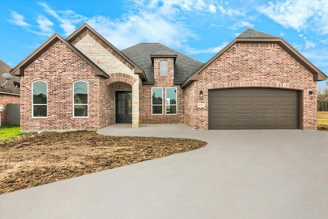 french provincial home featuring driveway, roof with shingles, stone siding, a garage, and brick siding