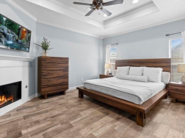bedroom with a tray ceiling, light wood-type flooring, ornamental molding, and a warm lit fireplace