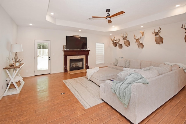 living room featuring ceiling fan, a raised ceiling, light hardwood / wood-style floors, and a tile fireplace