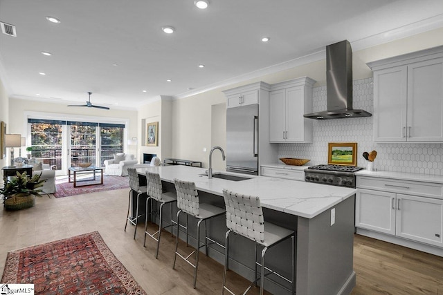 kitchen with stainless steel appliances, sink, wall chimney range hood, a center island with sink, and light hardwood / wood-style flooring